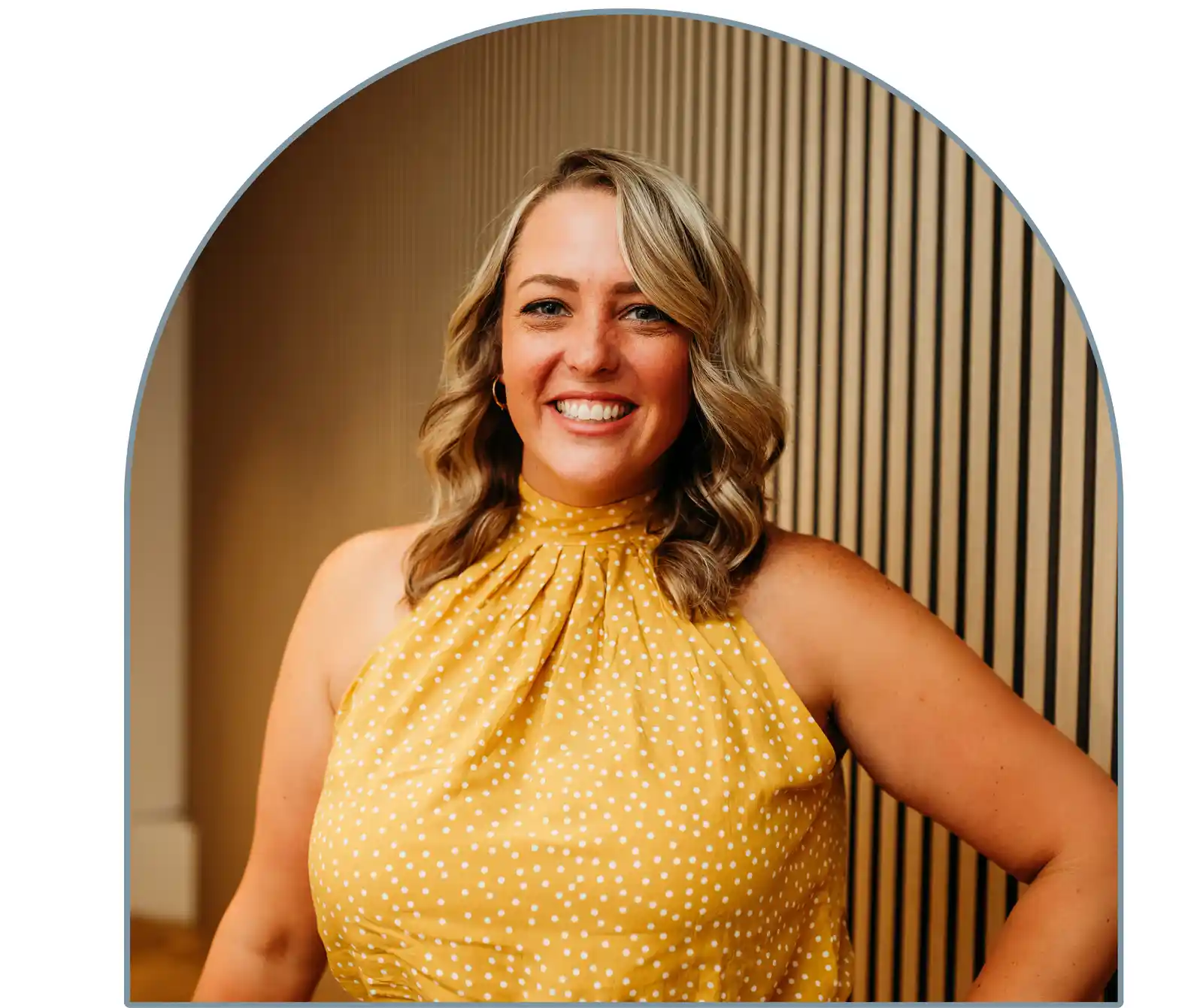 A woman with wavy hair is smiling, wearing a yellow polka-dot top. The image is framed with an arch-shaped border.