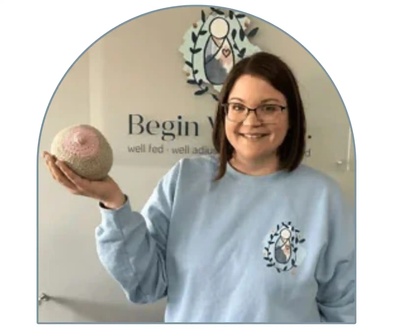 A woman with glasses is smiling and holding a crochet model of a breast. The text "Begin Well" is visible in the background, and the image is framed with an arch-shaped border.